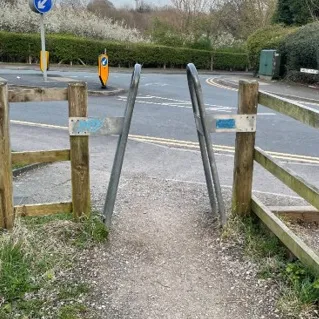 A metal, A-frame, motorised vehicle barrier, which prevents anyone wider than a human from going between a oath next to a road, and a track that leads off the path.