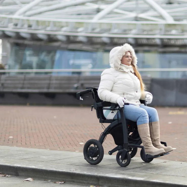 A lady in a wheelchair riding along a path, near the raised kerb, looking for a kerb drop.